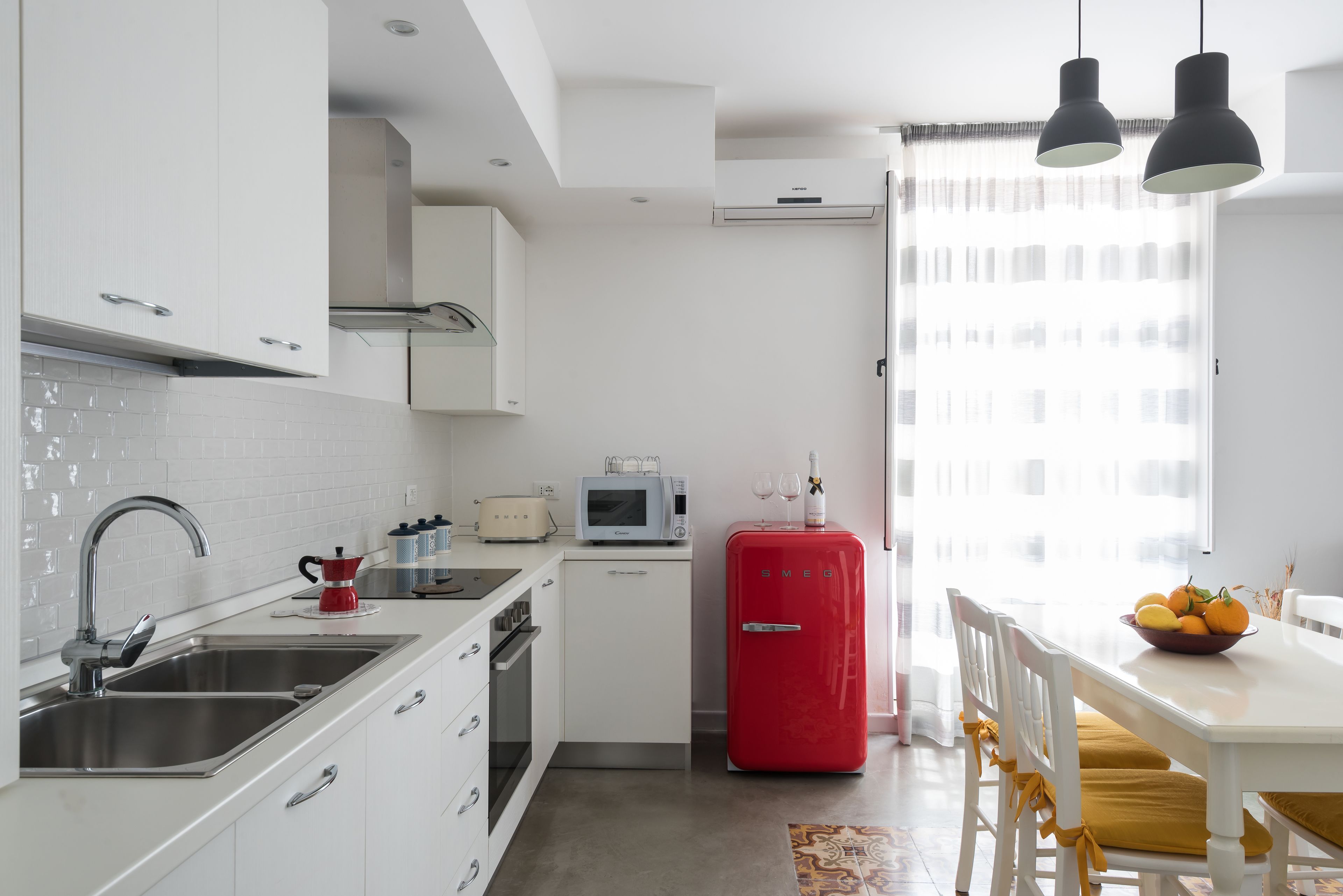 A modern kitchen with white cabinets, a stainless steel sink, a red refrigerator, and a dining area with a white table and yellow chairs.