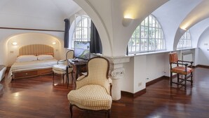 Ein geräumiges Zimmer mit einem großen Bett, einem Schreibtisch mit Computer und einem Stuhl. Große Fenster sorgen für viel Tageslicht.