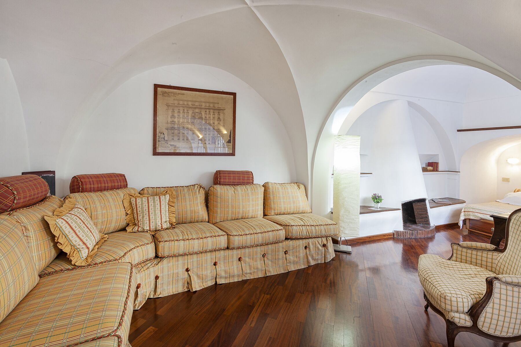 A spacious living room with a large, striped sofa set, wooden flooring, and a framed architectural drawing on the wall.