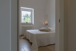 A bedroom with a bed, a window with a view of greenery, a radiator, and a bedside table with a lamp.