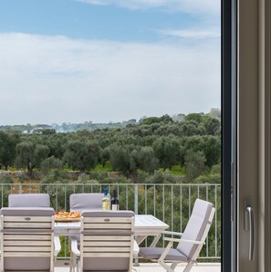 A balcony with a table and chairs, overlooking a landscape with olive trees and a stone wall.