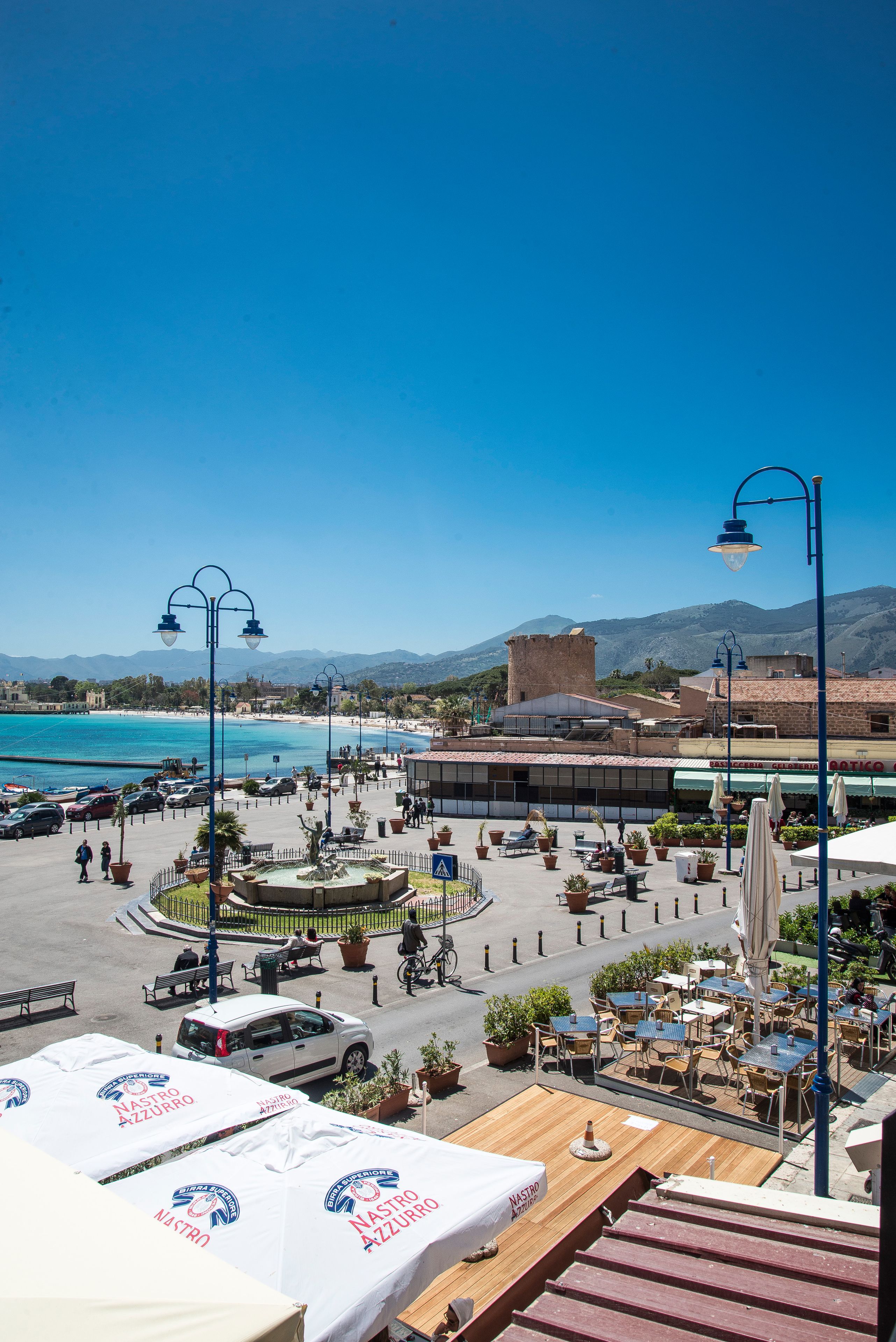 A coastal town with outdoor seating areas, a fountain, and a view of the sea and mountains.