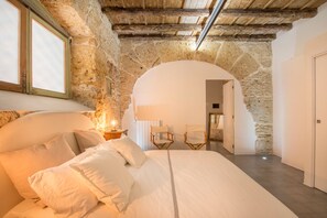 A bedroom with a stone archway, a bed with white linens, a nightstand with a lamp, and a wooden chair. - Stylish Home in Front of Casa Professa by Wonderful Italy (Palermo)