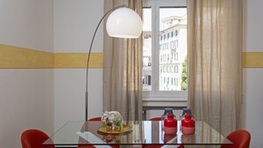 Una habitación de hotel moderna con una mesa de vidrio, sillas rojas, una lámpara blanca y vistas a edificios a través del ventanal.