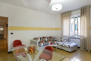 A modern living room with a glass coffee table, a sofa with patterned cushions, and a dining area with chairs.