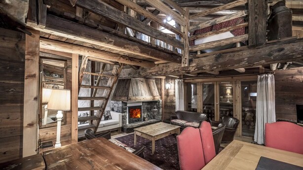 Interior rústico de una cabaña con chimenea de piedra, mobiliario de madera y una zona de dormir en entrepiso.