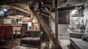 Un interior rústico con vigas de madera a la vista, una pared de piedra, una escalera de madera, un sofá de cuero y un comedor con mobiliario de madera.