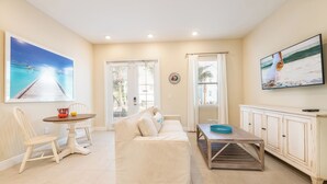 A living room with a beige sofa, a dining table with chairs, a TV, and a large window with curtains. - Livin' for the Weekend Cottage near Disney | Resort & Waterpark Access - 8021FF (Kissimmee)