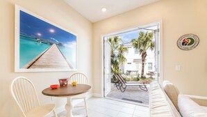Une pièce comprenant une table à manger, des chaises, un canapé, un grand tableau représentant une jetée, et une vue sur des palmiers et une maison à l’extérieur.
