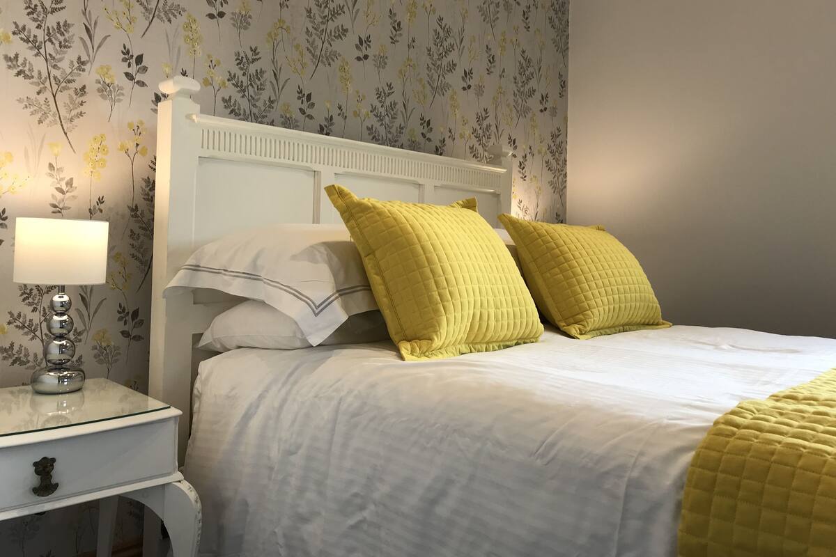 Un lit bien fait, avec une literie blanche et deux coussins jaunes, une table de chevet avec une lampe et un papier peint à motifs floraux.