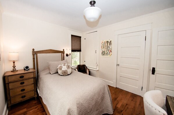 A bedroom with a wooden four-poster bed, a nightstand with a lamp, a chair, and a door with a decorative panel.