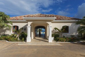 A Mediterranean-style building with a tiled roof, an arched entrance, and a landscaped garden with palm trees and red flowers.