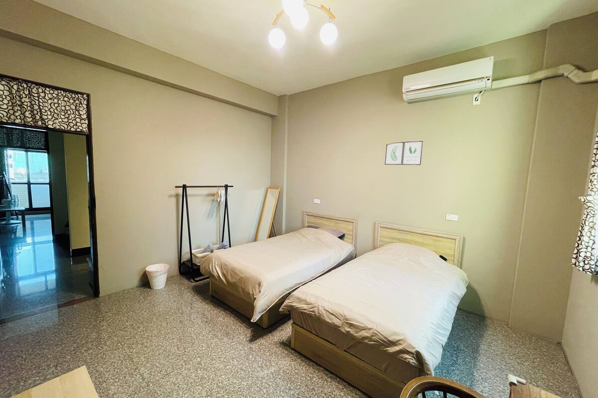 Habitación de hotel con dos camas individuales, una silla de madera, una mesita pequeña, un equipo de aire acondicionado y un espejo con marco decorativo.