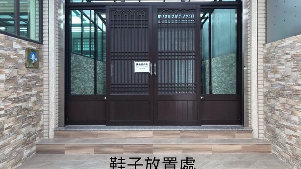 L'ingresso di un edificio con una porta in legno scuro, un cartello che invita a non indossare scarpe all'interno e una parete con effetto pietra su entrambi i lati.