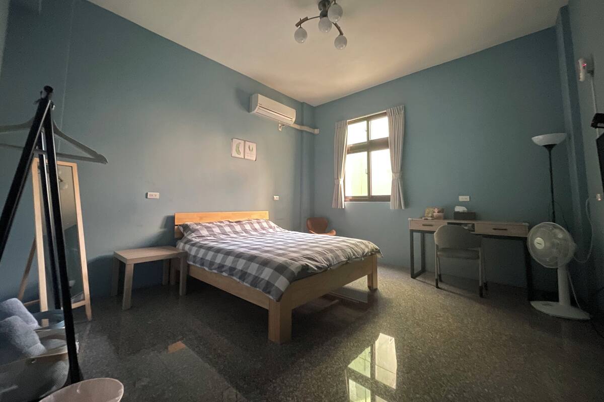 Un dormitorio con cama, escritorio, silla, ventilador de pie y ventana con cortinas.