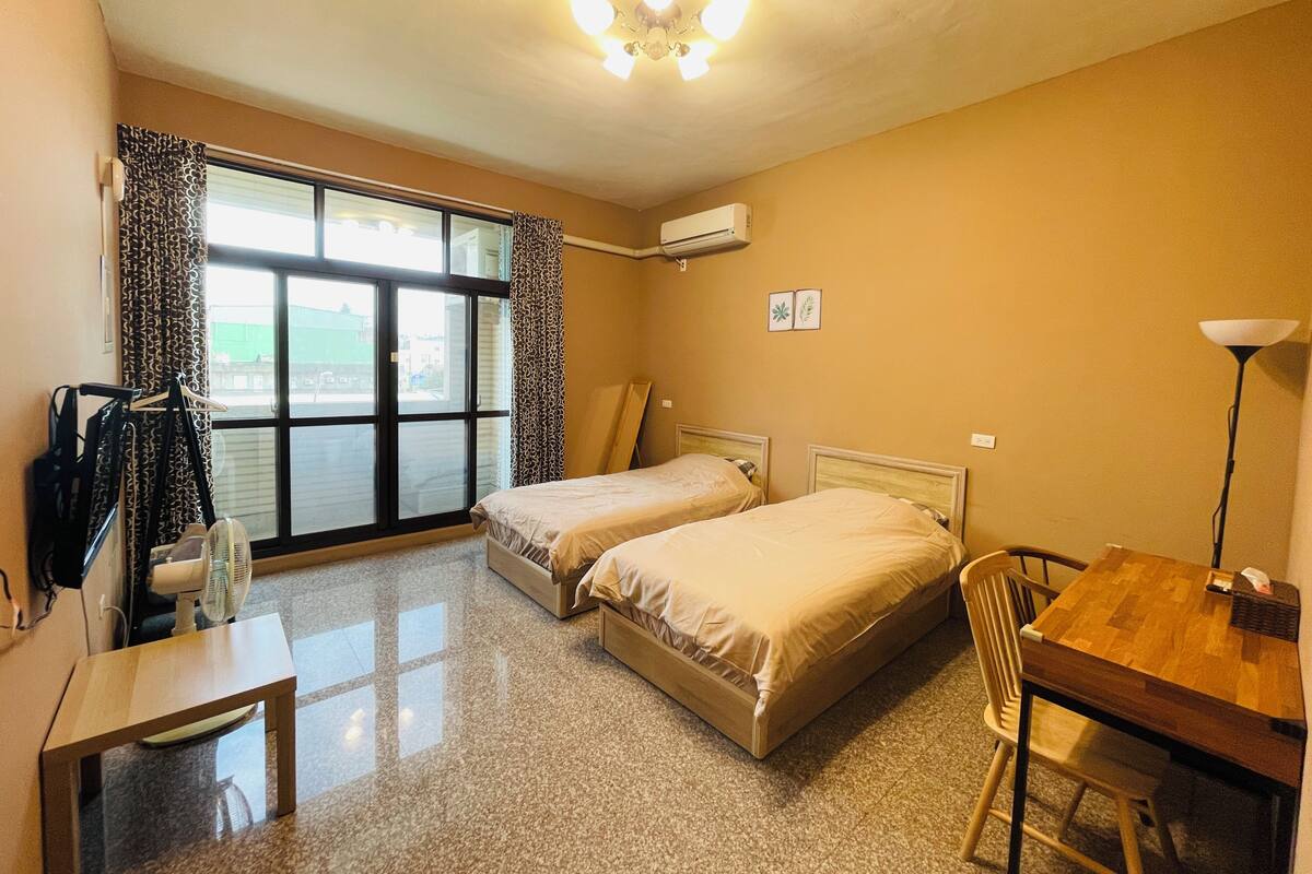 Habitación de hotel con dos camas, un escritorio, una silla, una lámpara y vistas a través de un ventanal grande.