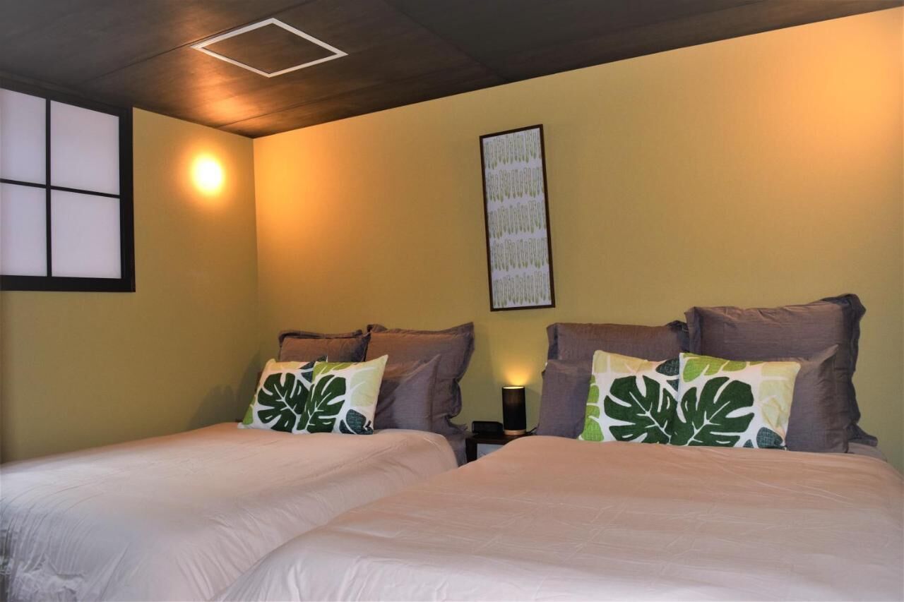 Habitación de hotel con dos camas, cada una con un almohadón estampado con hojas. Se aprecia una lámpara de pared e imagen en un marco colgada en la pared.