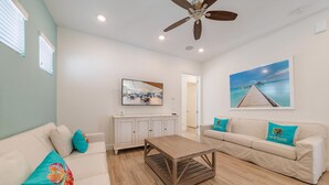 A living room with a wooden coffee table, two sofas with teal pillows, a TV on a cabinet, and a ceiling fan. - Tropical Oasis Cottage | Hot Tub + Margaritaville & Waterpark Access - 3021LA (Kissimmee)