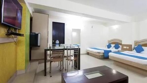 A hotel room with a dining table, chairs, a sofa with blue pillows, a TV mounted on the wall, and a water dispenser. - Hotel Metro Express (New Delhi)