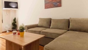A living room with a beige sofa, a wooden coffee table, a small potted plant, a TV, and a painting of a sunset. - Dervenion-Athens Cozy Apartment (Athens)