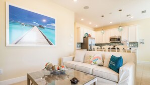 A living room with a glass coffee table, a beige sofa, and a kitchen in the background.