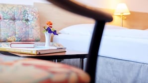 A hotel room with a bed, a bedside table with a lamp, a floral-patterned pillow, and a small vase with flowers. - Ring Hotel (Wiesbaden)