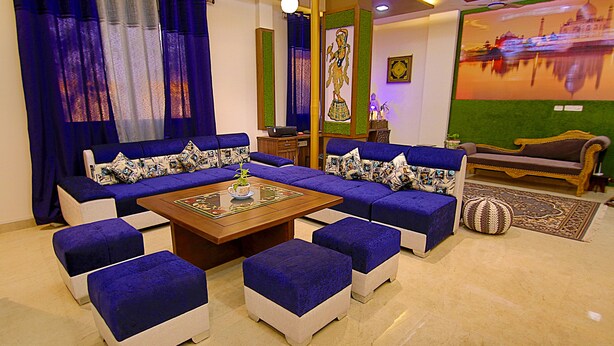 A living room with a sectional sofa, ottomans, a coffee table, and a television screen displaying a cityscape. - The Hideout Agra Homestay (Agra)