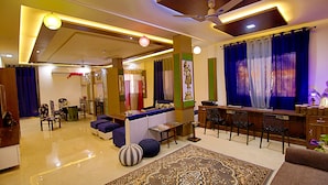 A spacious living area with a sofa, a coffee table, and a dining table. There is a ceiling fan and a chandelier. - The Hideout Agra Homestay (Agra)