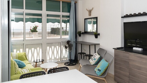 Un salon moderne avec un canapé, une table à manger et un téléviseur. Il y a des fenêtres donnant sur la mer et un balcon agrémenté de plantes.