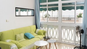 Une pièce avec un canapé vert, deux petites tables rondes, un ventilateur et une vue sur la plage grâce à de grandes fenêtres.