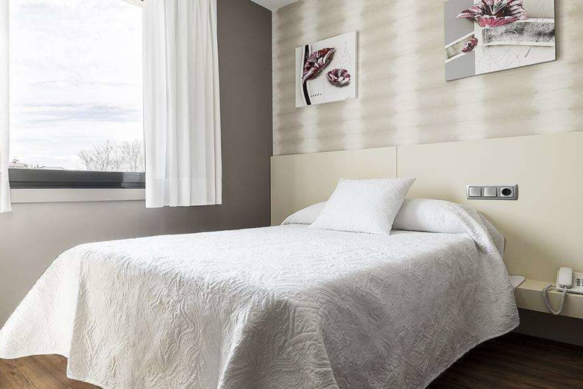 A bedroom with a bed, a bedside table, a nightstand, a window with curtains, and wall art.