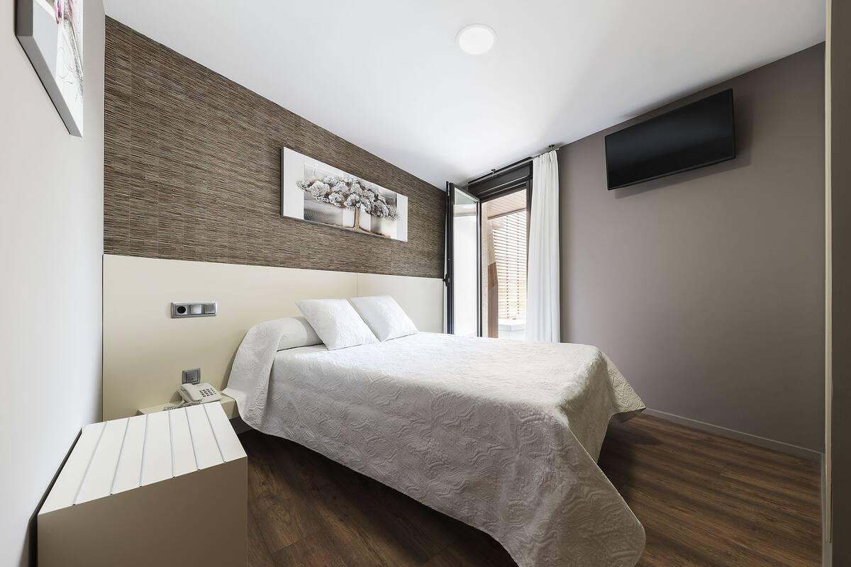 A modern bedroom with a bed, a flat-screen TV, a painting, and a window with blinds.