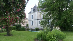 Un edificio grande e histórico con fachada simétrica, rodeado por un jardín bien cuidado con diversas plantas y un árbol de flores rosadas.