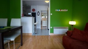 Sala de estar com parede verde, sofá vermelho, mesa de jantar com cadeiras e cozinha visível por uma porta aberta.