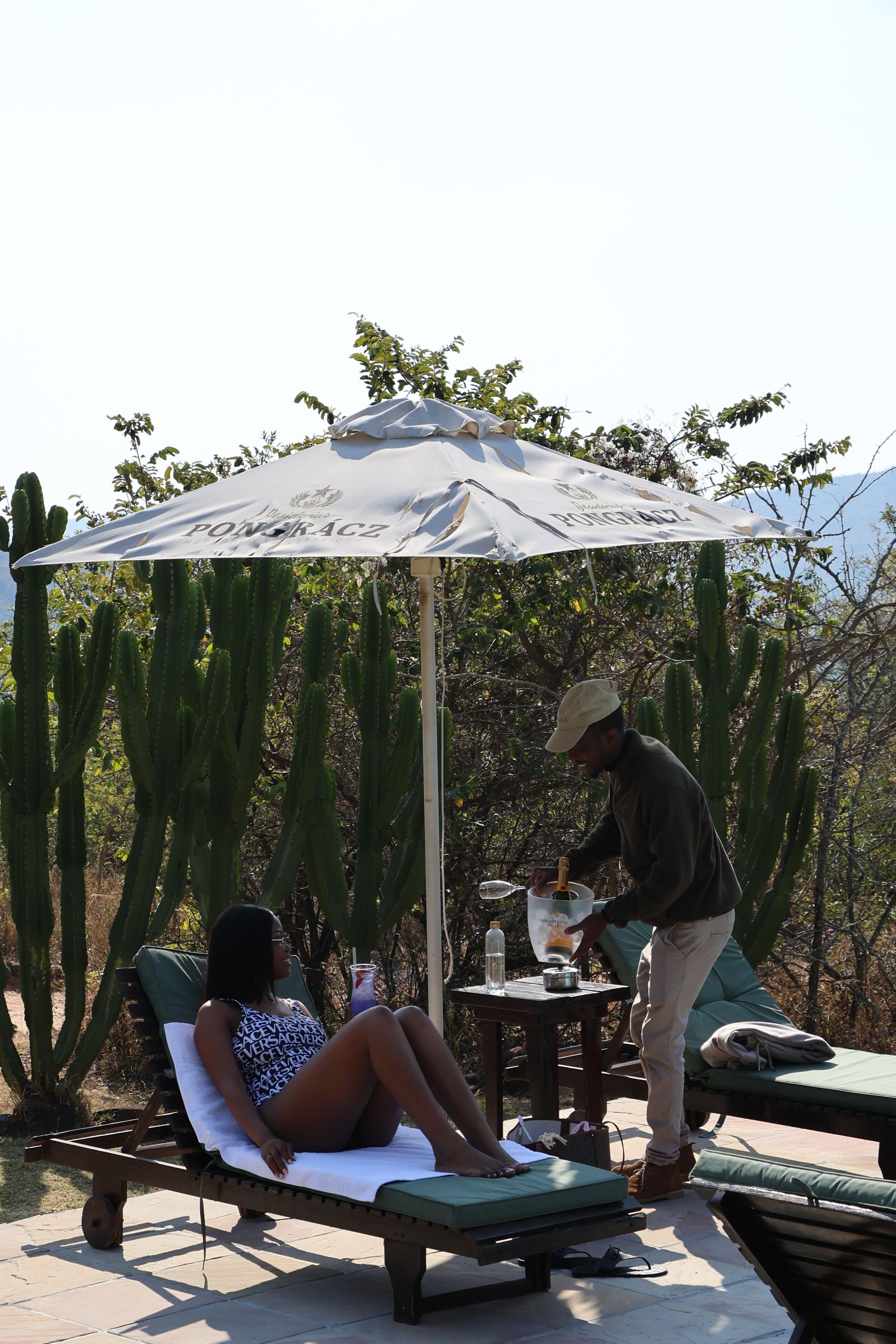Une personne sert une boisson à une autre personne assise sous un parasol, dans un décor désertique parsemé de cactus.