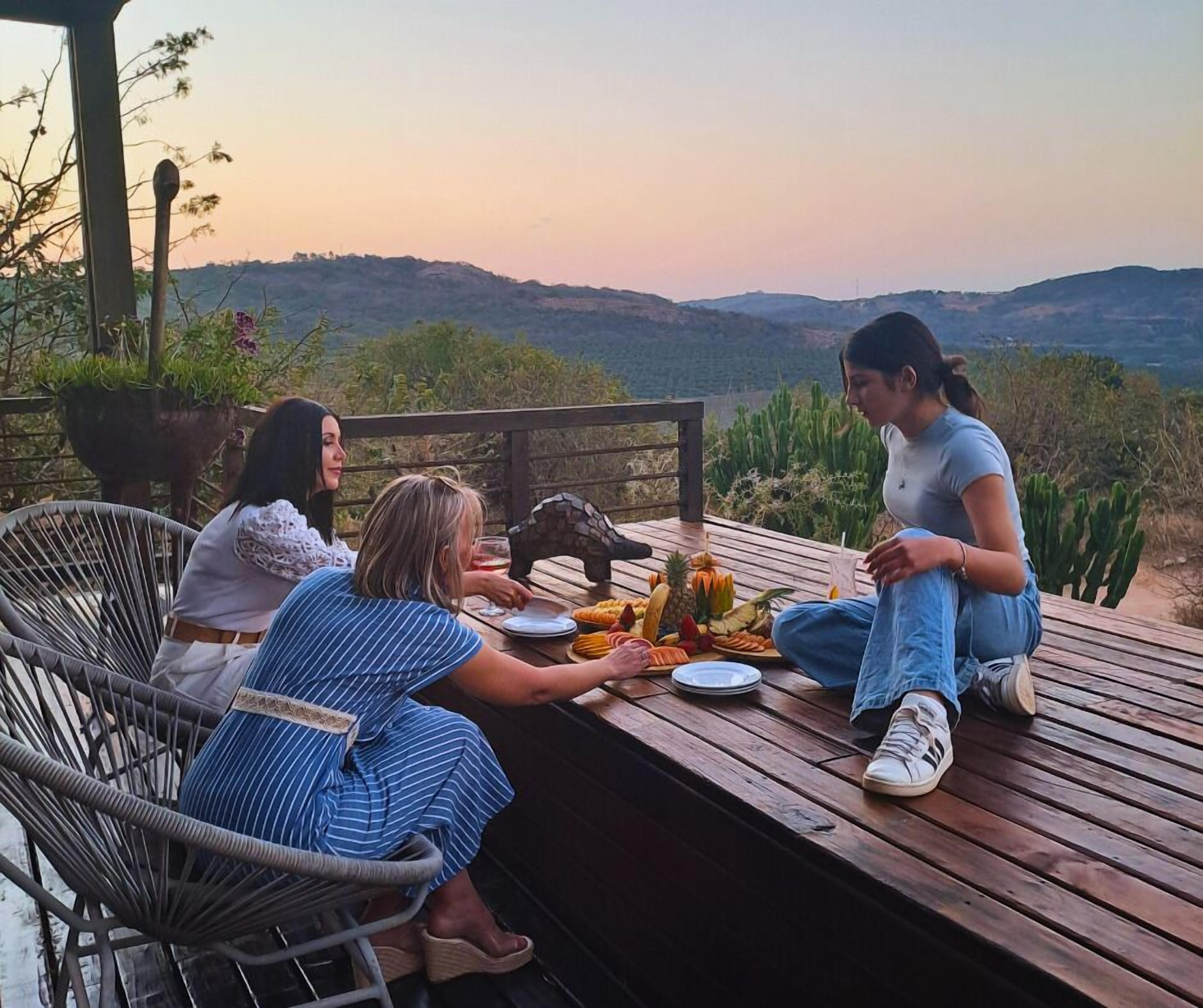 Una terraza de madera con una mesa dispuesta para una comida, rodeada de sillas, con vistas a un paisaje montañoso.