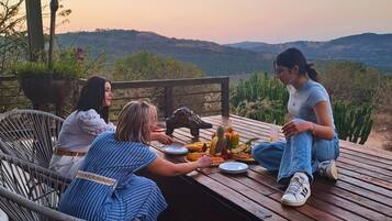 Una terraza de madera con una mesa dispuesta para una comida, rodeada de sillas, con vistas a un paisaje montañoso.