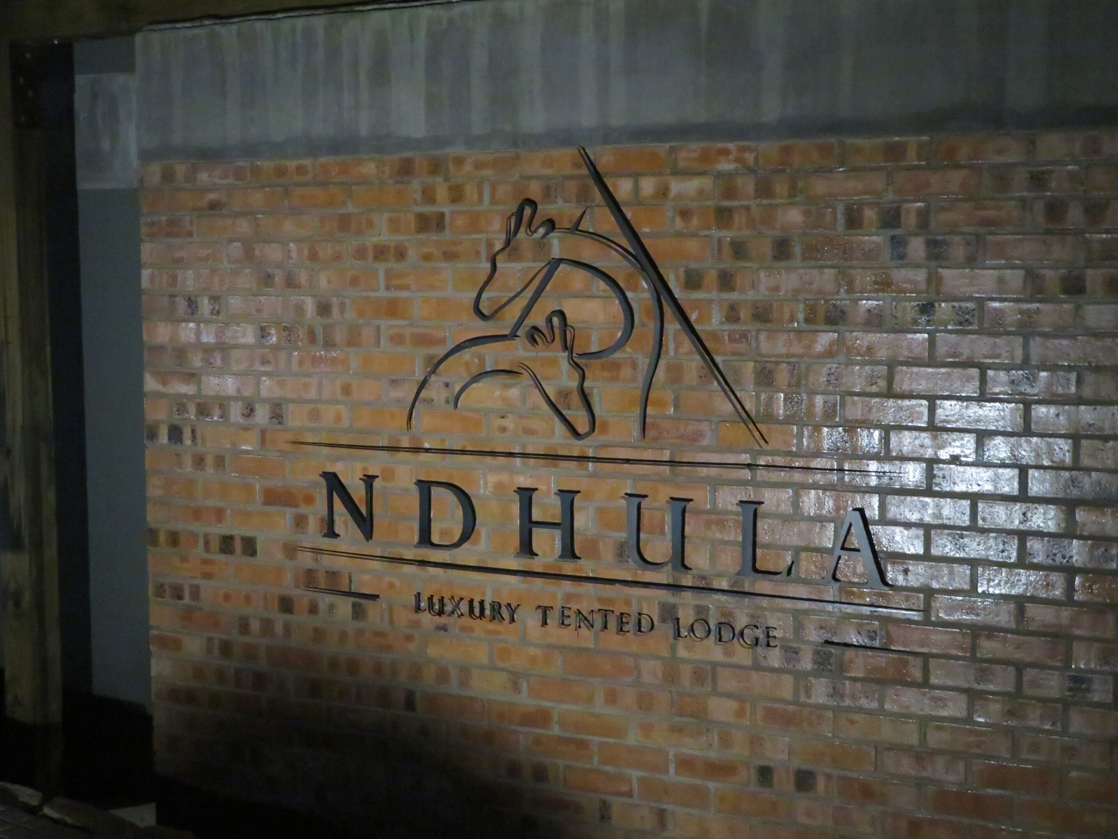 A brick wall with the logo and name "NDHULA LUXURY TENTED LODGE" and a stylized horse silhouette.