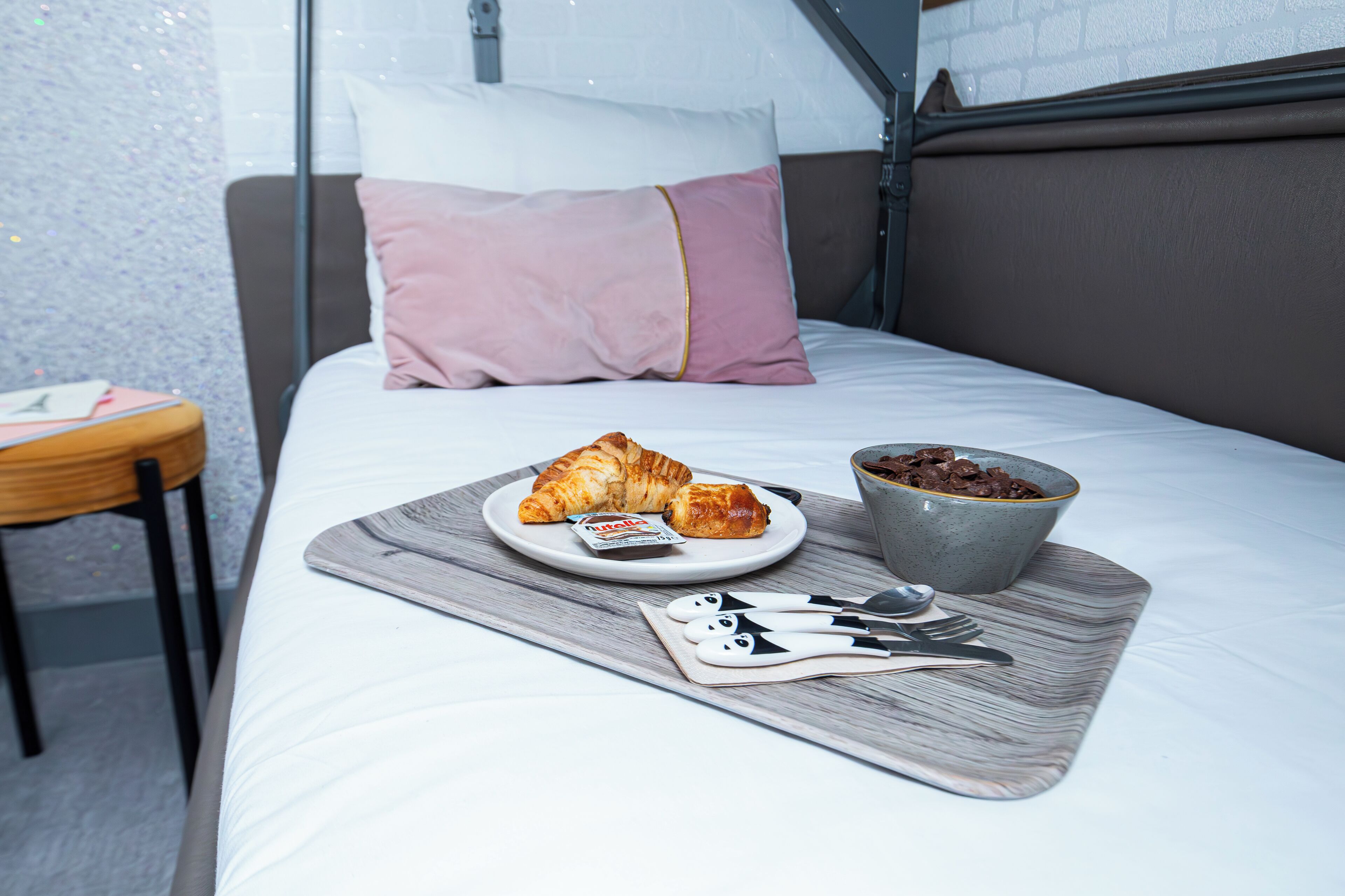 A neatly made bed with a pillow, a plate with croissants and a bowl of cereal, and a tray with cutlery.