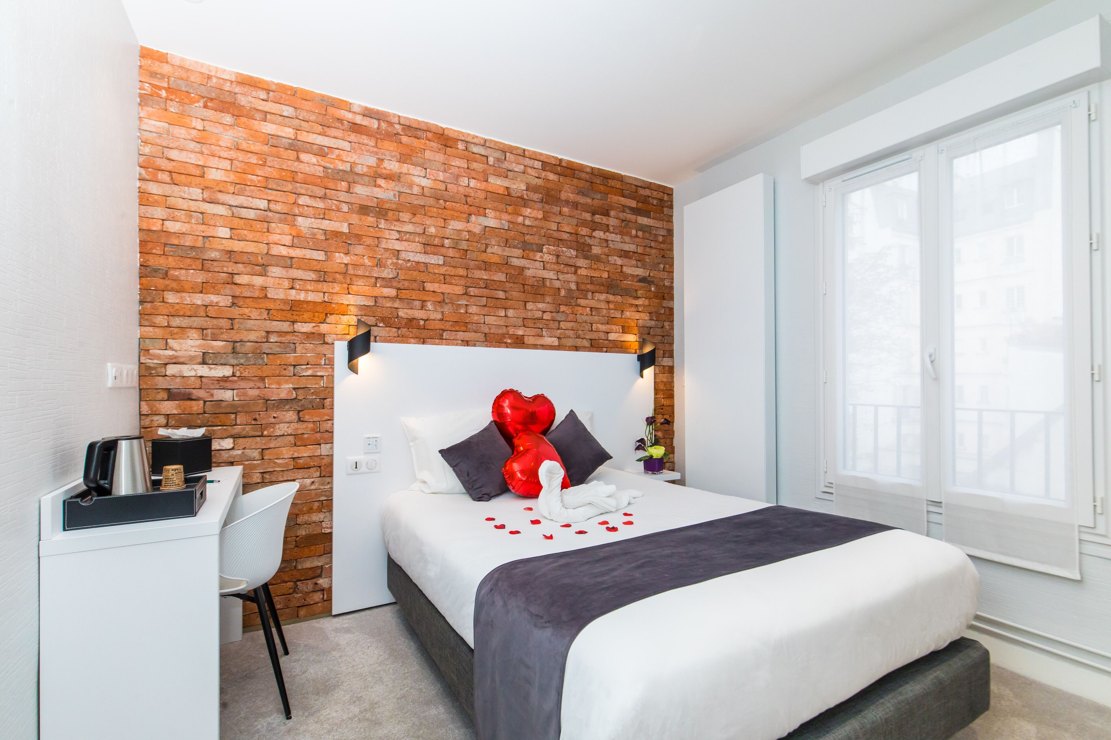 Una habitación de hotel con una pared de ladrillo decorativa, una cama con colcha blanco y gris, un escritorio con tetera y una silla.