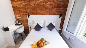Une chambre d’hôtel avec un mur en briques, un lit blanc avec des oreillers noirs, un bureau avec une chaise et un plateau de petit-déjeuner posé sur le lit.