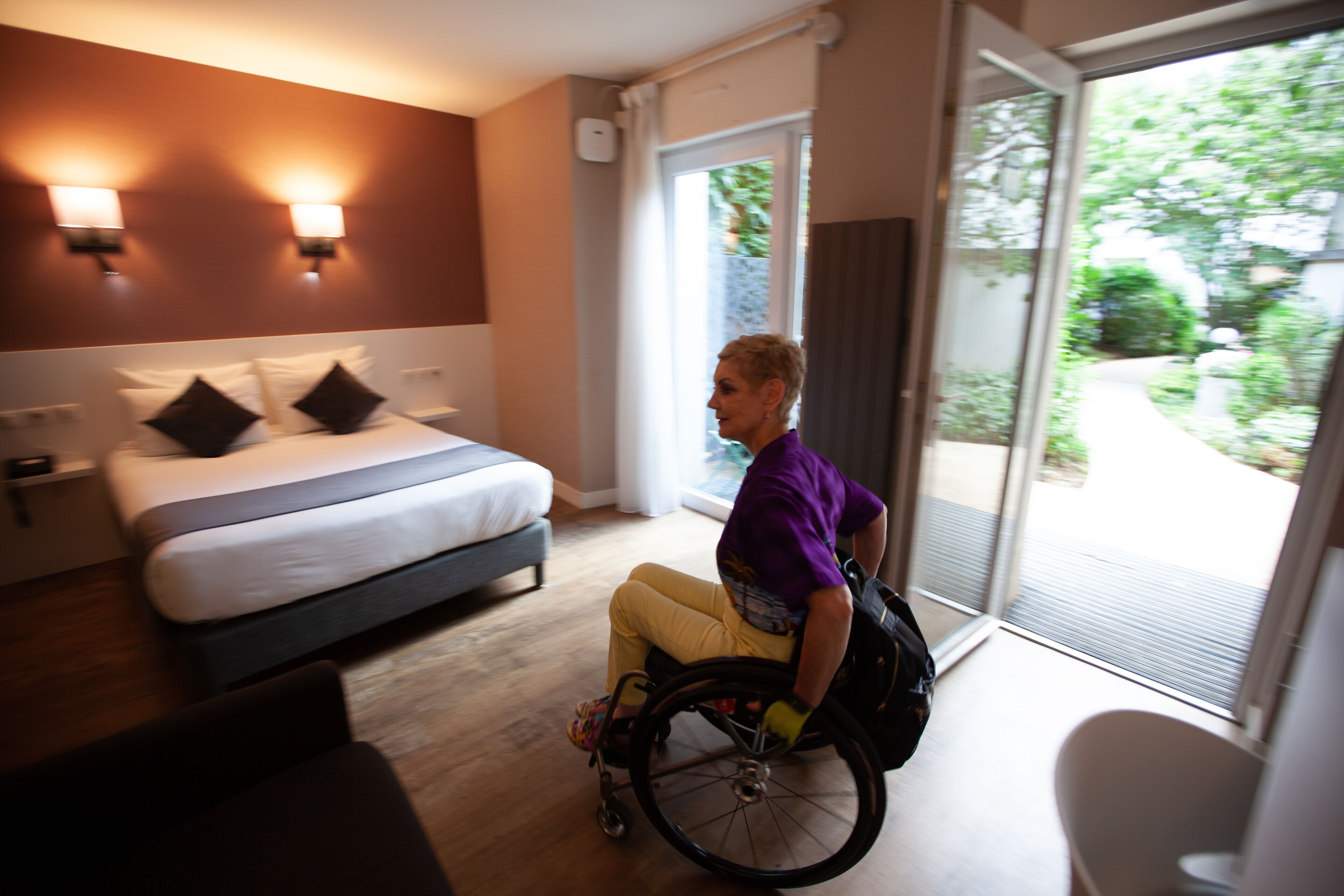 Habitación de hotel con cama, silla de ruedas y vistas al exterior.