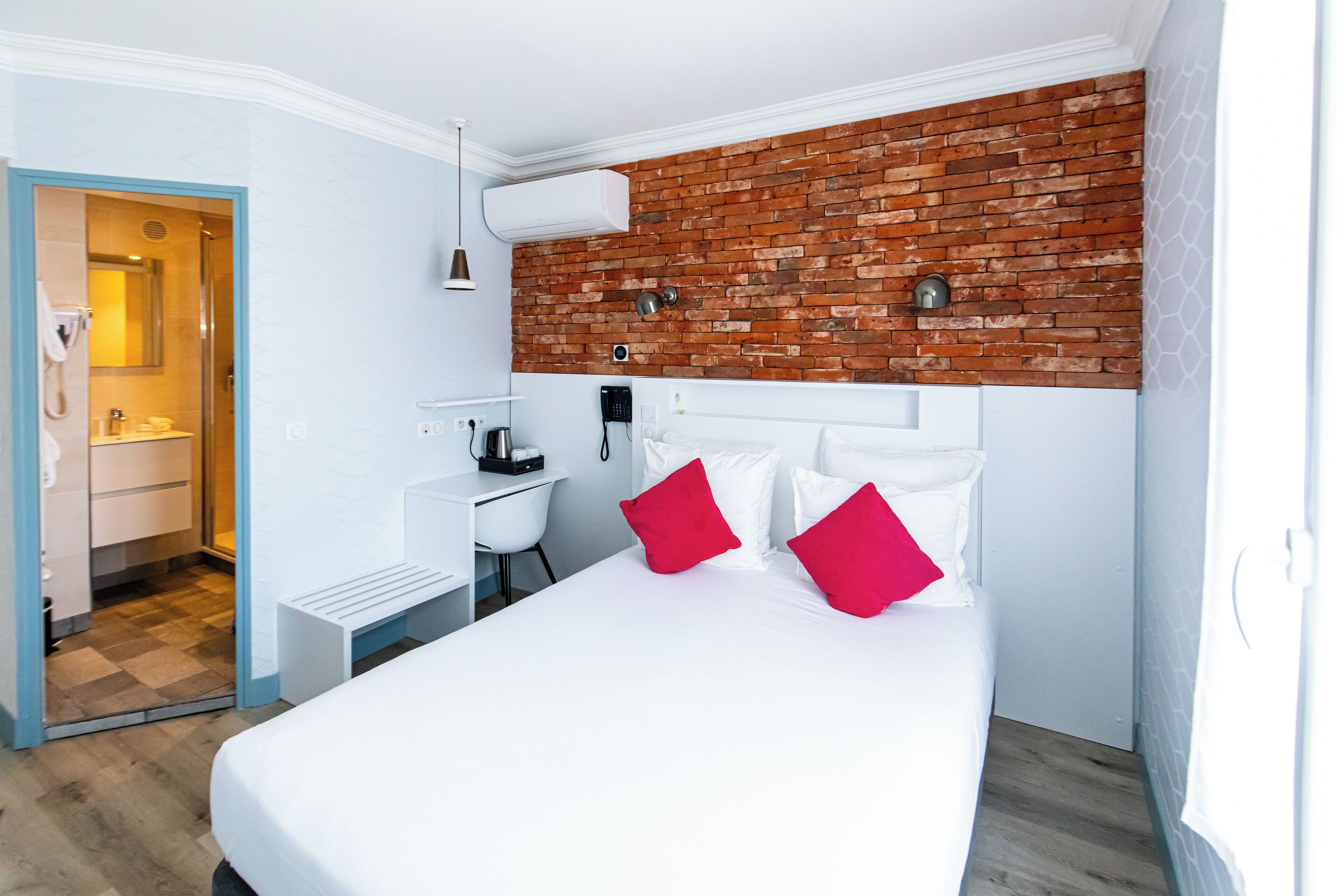 A hotel room with a white bed, red pillows, a brick accent wall, a desk with a chair, and a bathroom visible through an open door.