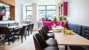 Un espace repas moderne avec des banquettes en cuir noir et une longue table, une cuisine lumineuse avec des meubles roses et de grandes fenêtres qui laissent entrer la lumière naturelle.