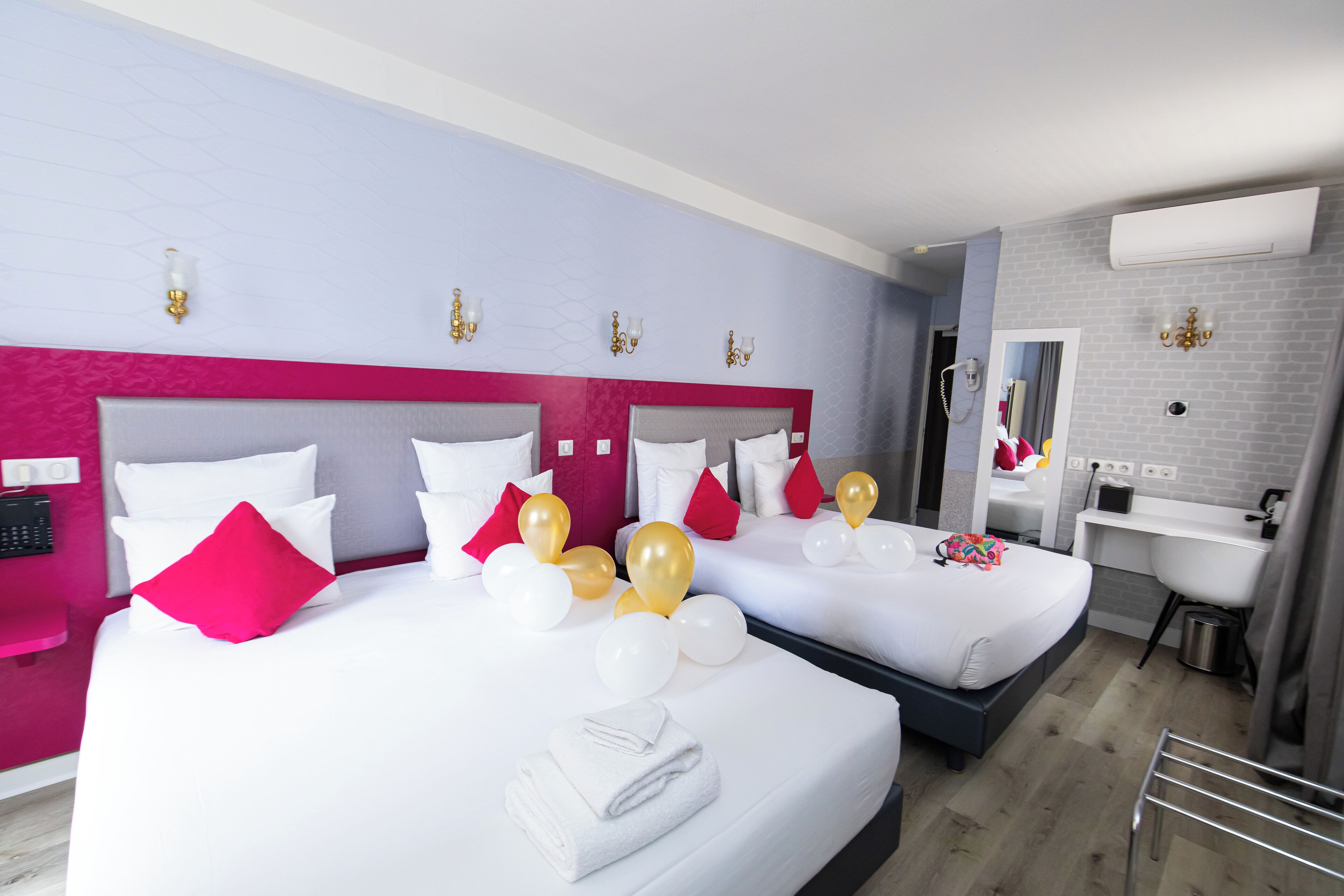 A hotel room with two beds, each with white linens and red and gold balloons. There is a bathroom with a sink and mirror.