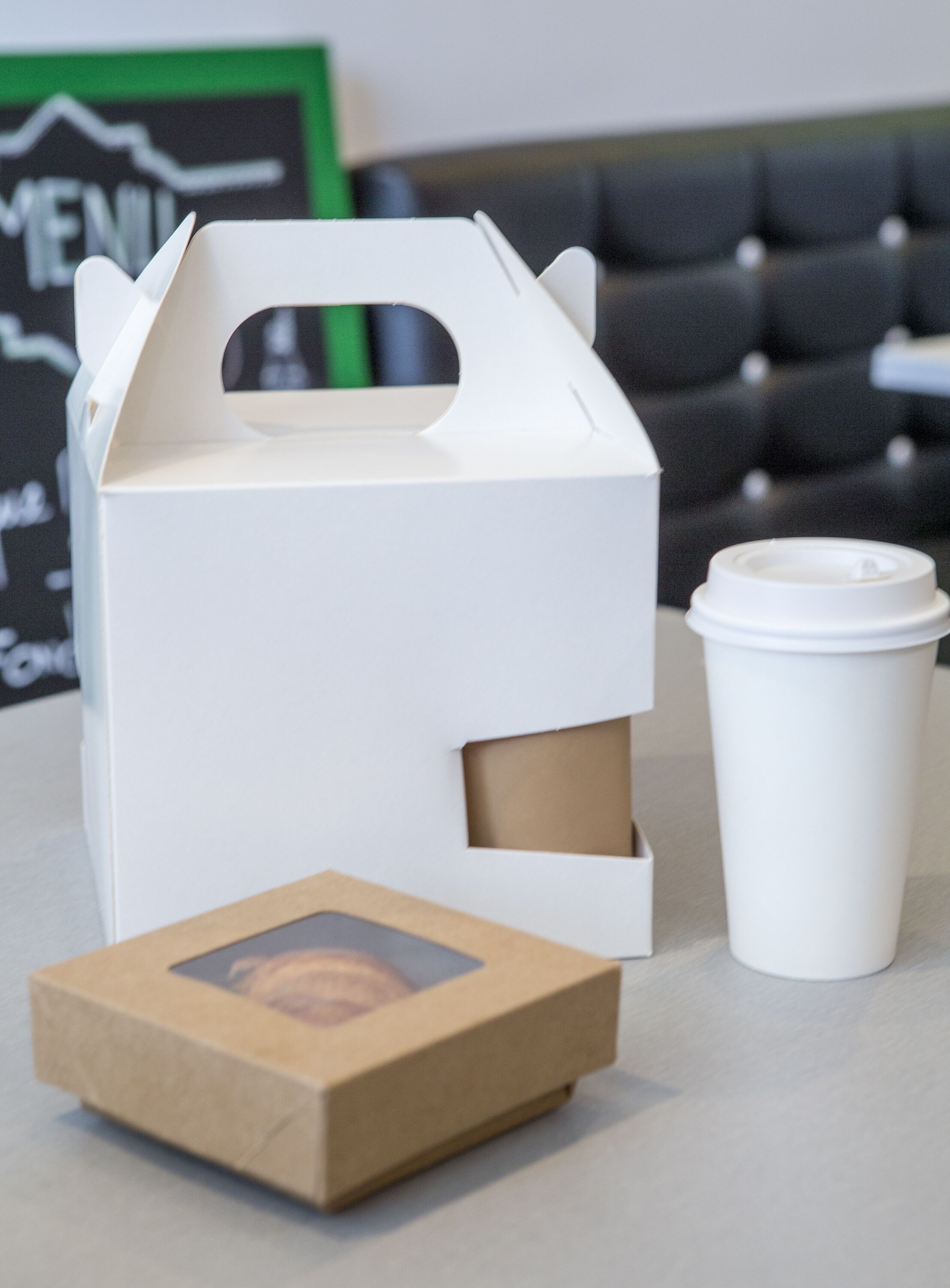 Un recipiente blanco para comida para llevar con asa, una taza desechable blanca y una caja de cartón pequeña con una imagen sobre una mesa.