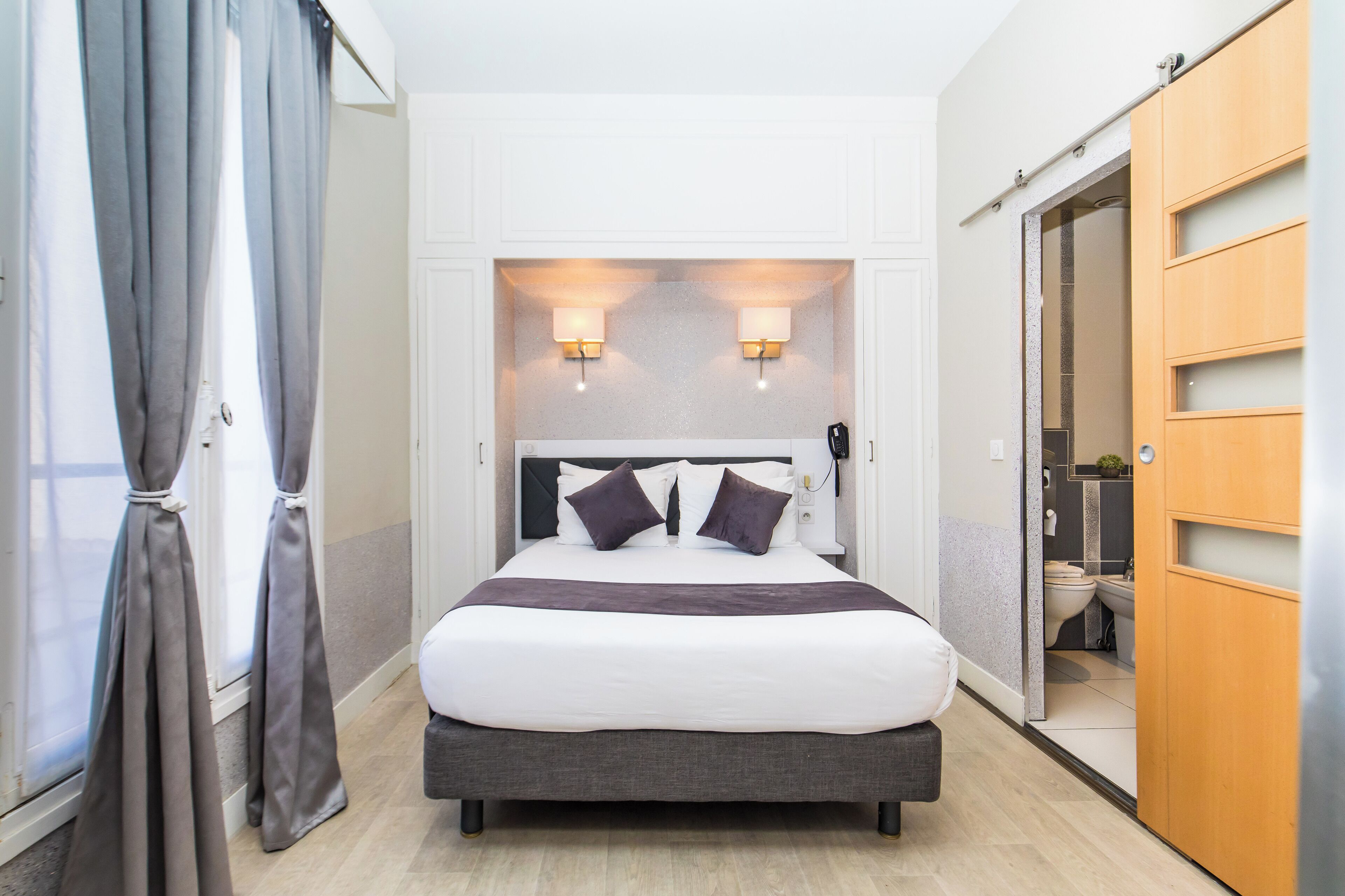 A hotel room with a large bed, two bedside lamps, a wardrobe, and a bathroom visible through an open door.