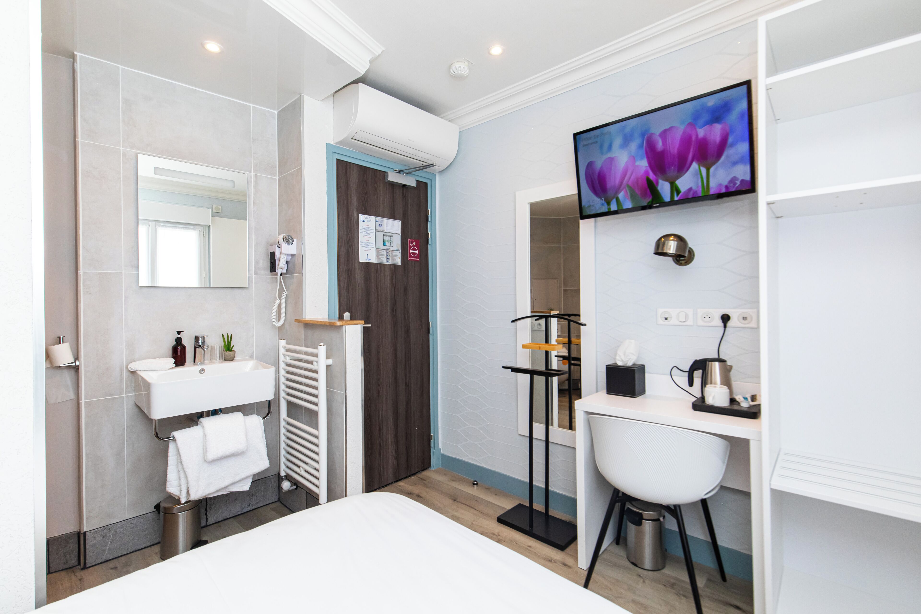 Un baño moderno con lavamanos, espejo e inodoro. Hay un televisor montado en la pared y un escritorio pequeño con una silla.