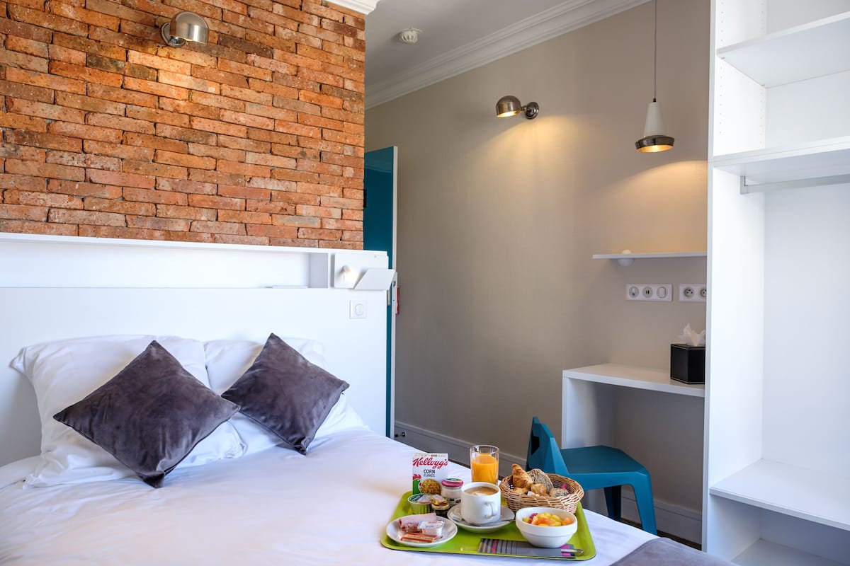 Une chambre d’hôtel avec un mur en briques, un lit blanc, deux oreillers et un plateau de petit-déjeuner comprenant du café, des croissants et des fruits.