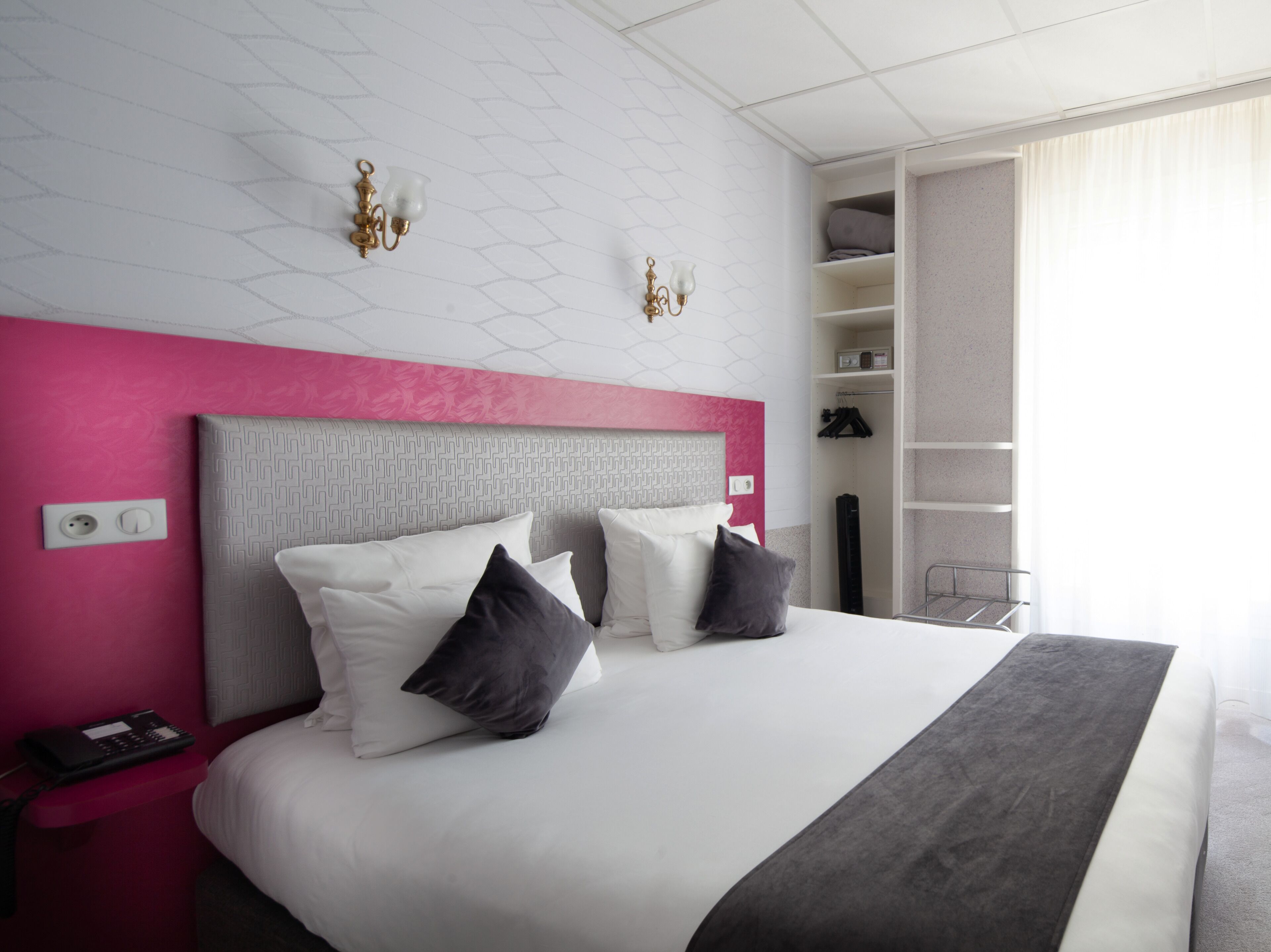 Una habitación de hotel moderna con una cama grande, una pared de acento rosa, un cabecero blanco y un armario empotrado con estantes.